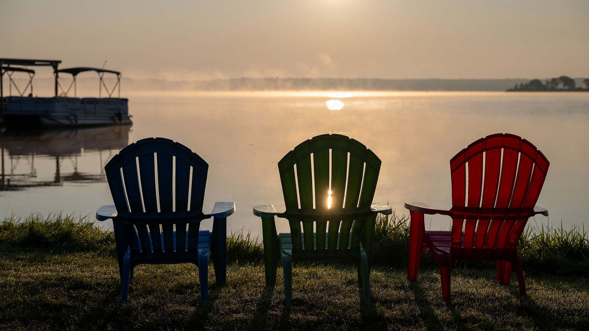 Three vacant, multi-color lawn chairs looking out over a steaming lake at sunset